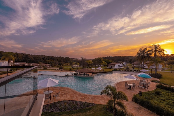 A swimming pool with a sunken area, surrounded by outdoor seating and umbrellas, with a view of a residential area in the background.