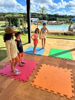 Um grupo de crianças em cima de tapetes de espuma interligados ao ar livre, com uma piscina e área coberta ao fundo.
