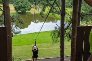 A person on a zip line over a body of water, with a grassy area and trees in the background.