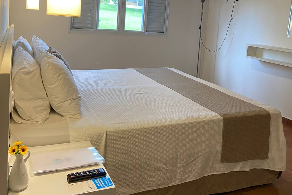 Une chambre à coucher comprenant un lit, une table de chevet, une lampe, un téléviseur et une fenêtre avec des stores.