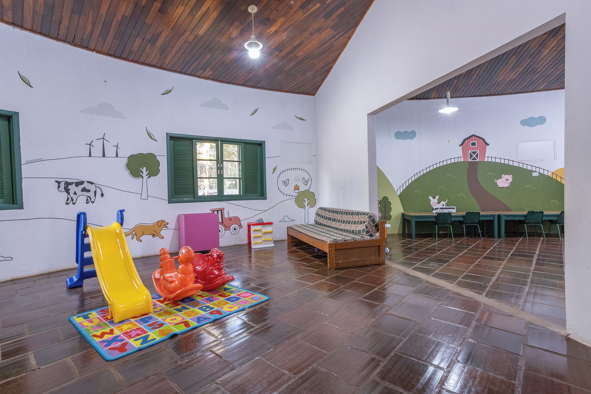 A children's playroom with a wooden floor, a play mat, colorful toys, a bench, and a mural depicting farm scenes.