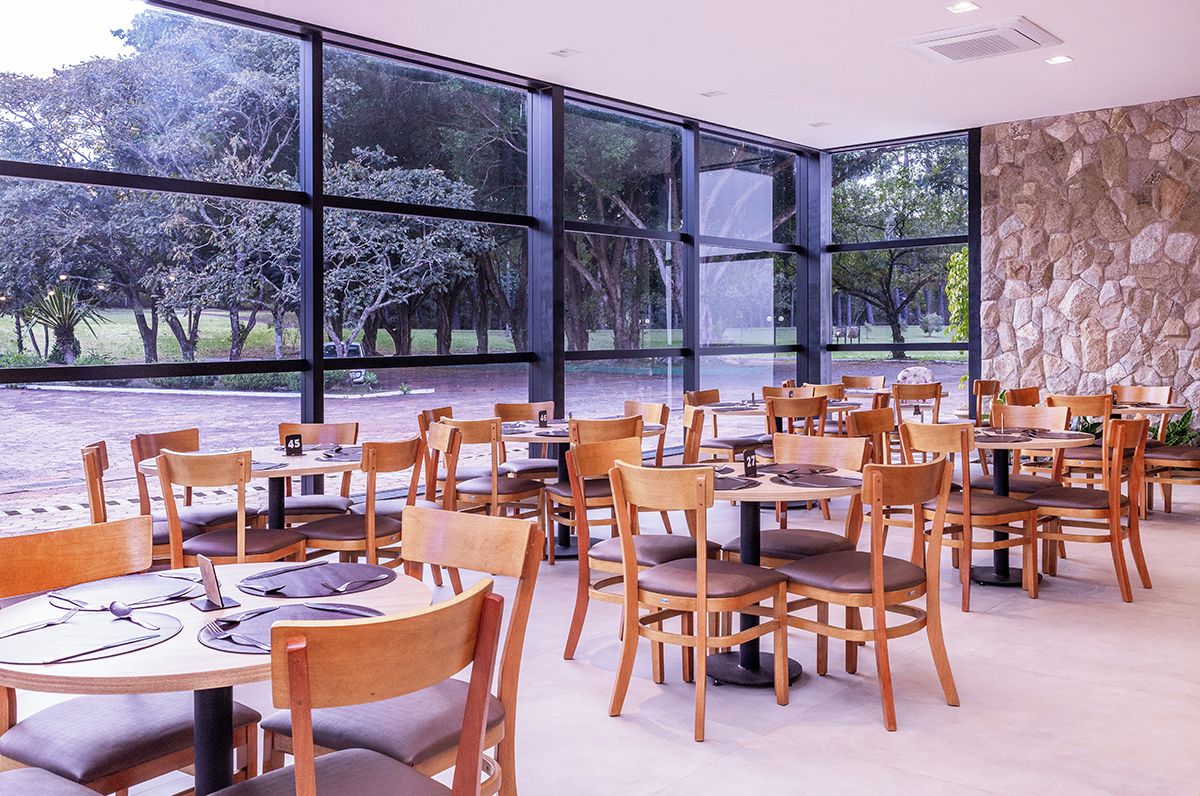 A restaurant with large windows offering a view of trees and a body of water. Tables are arranged with chairs, and the interior has a stone accent wall.