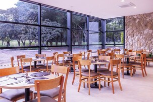 Un restaurante con grandes ventanales que ofrecen vistas a árboles y un cuerpo de agua. Las mesas están dispuestas con sillas y el interior cuenta con una pared de acento de piedra.