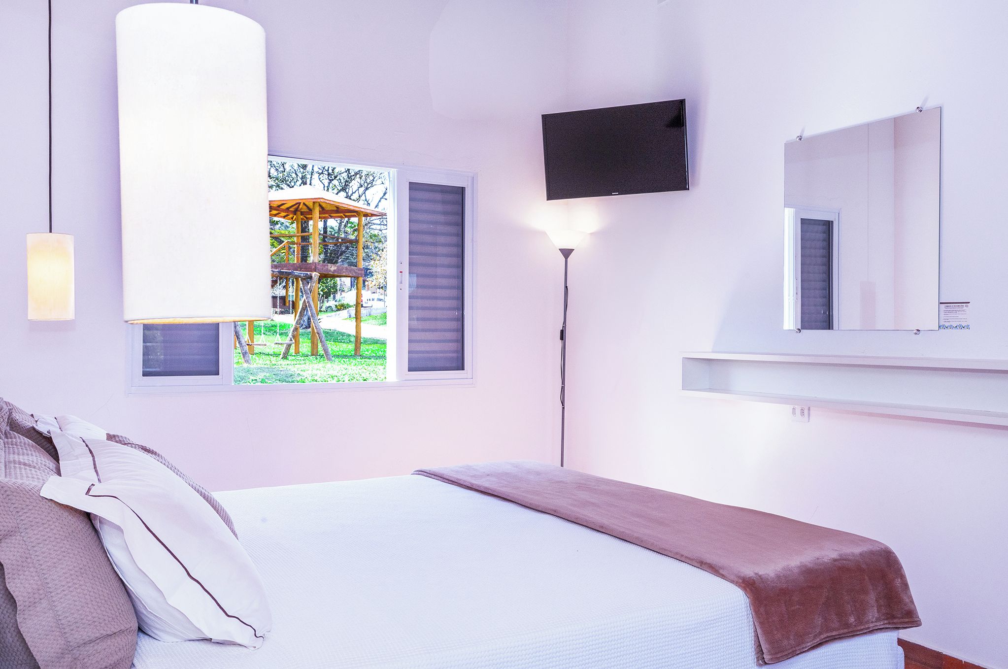 A bedroom with a bed, a window with a view of a garden, a mounted television, and a bedside lamp.