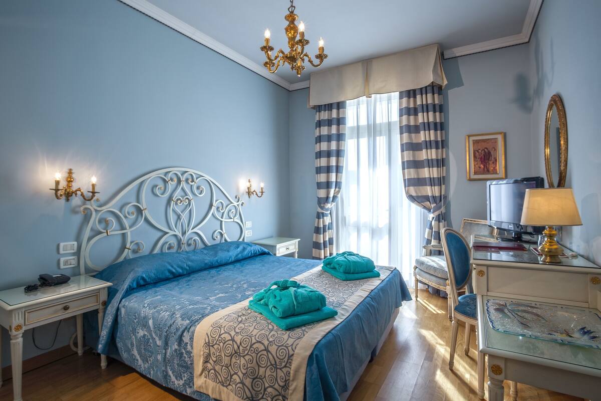 Een slaapkamer met een bed, nachtkastjes, een bureau, een stoel, een kroonluchter en een raam met gordijnen.