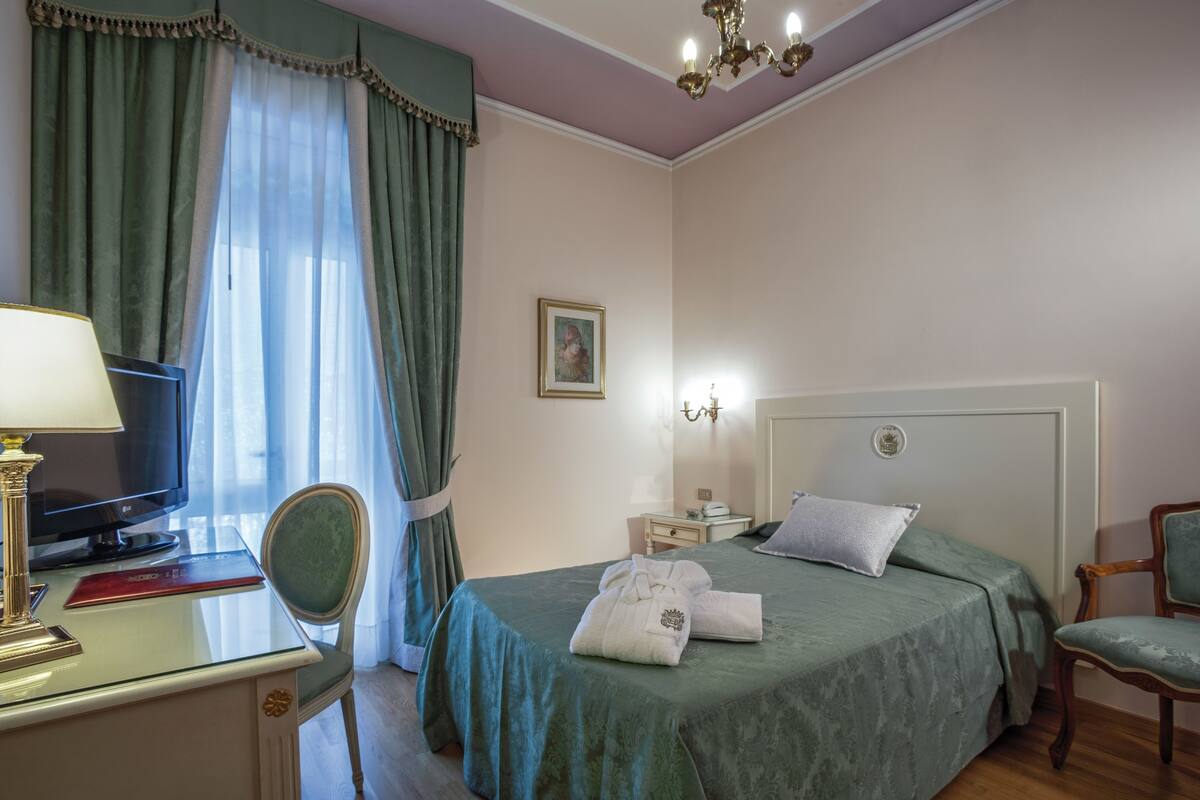 Een hotelkamer met een bed, een bureau met lamp, een stoel, een nachtkastje en een foto aan de muur.