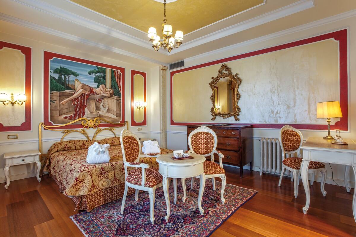 Een klassieke kamer met een bed, twee stoelen, een tafeltje, een spiegel en een radiator.