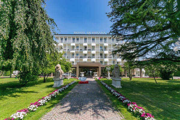 A hotel with a central entrance, surrounded by a garden with a paved pathway and flower beds. - Hotel Quisisana (Abano Terme)