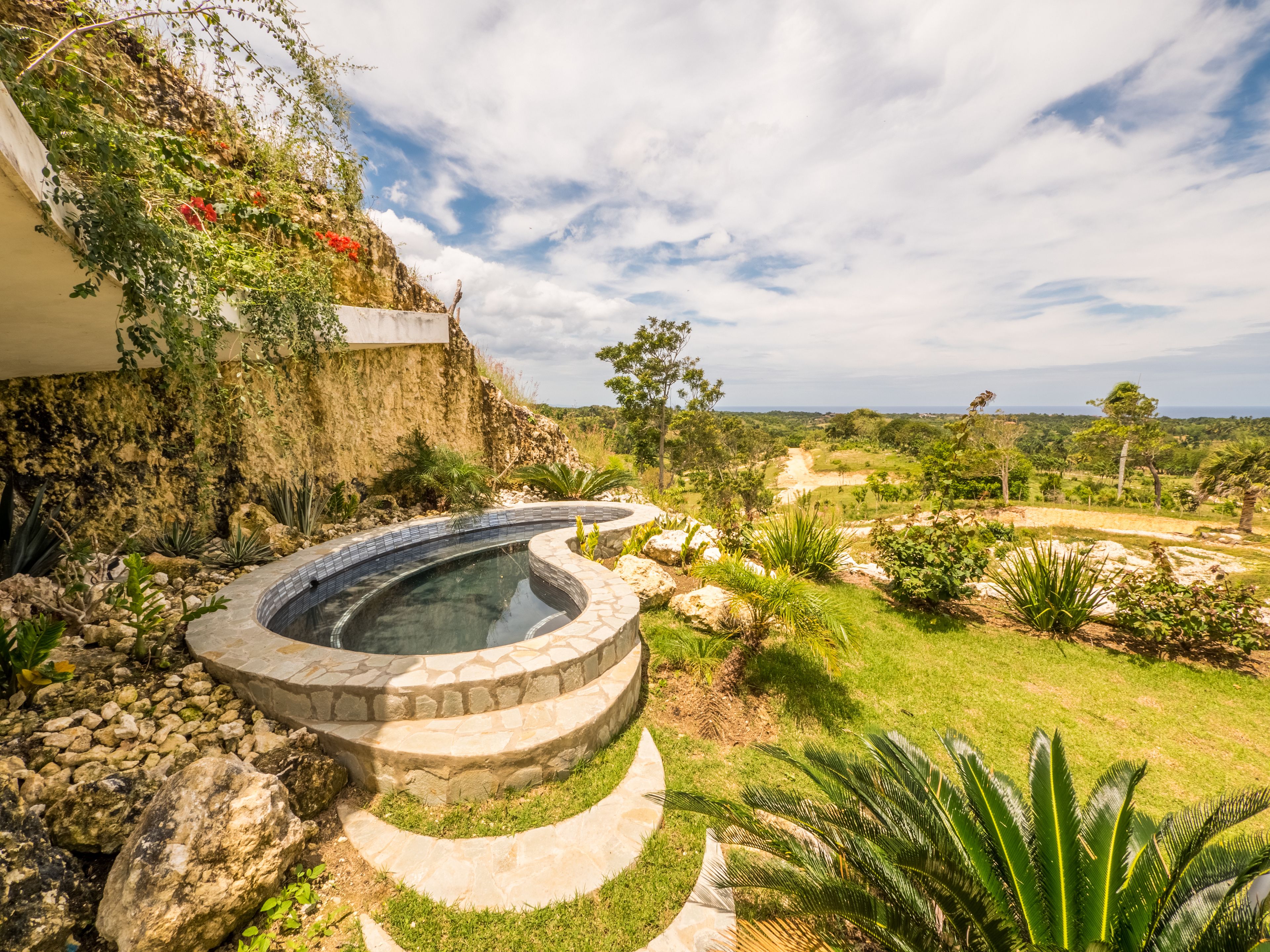 A hot tub with a stone design, surrounded by a garden with various plants and a view of a hilly landscape.