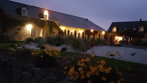Un bâtiment en pierre doté d’un toit en pente, illuminé par des lumières chaleureuses, entouré d’un jardin où poussent des fleurs jaunes et dont le sol est recouvert de neige.
