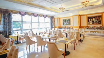 A spacious dining area with multiple tables and chairs, large windows with curtains, and chandeliers.