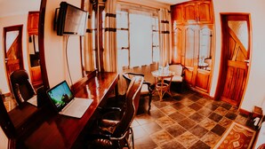 Quarto com escrivaninha de madeira, cadeiras, televisão e janela com cortinas.