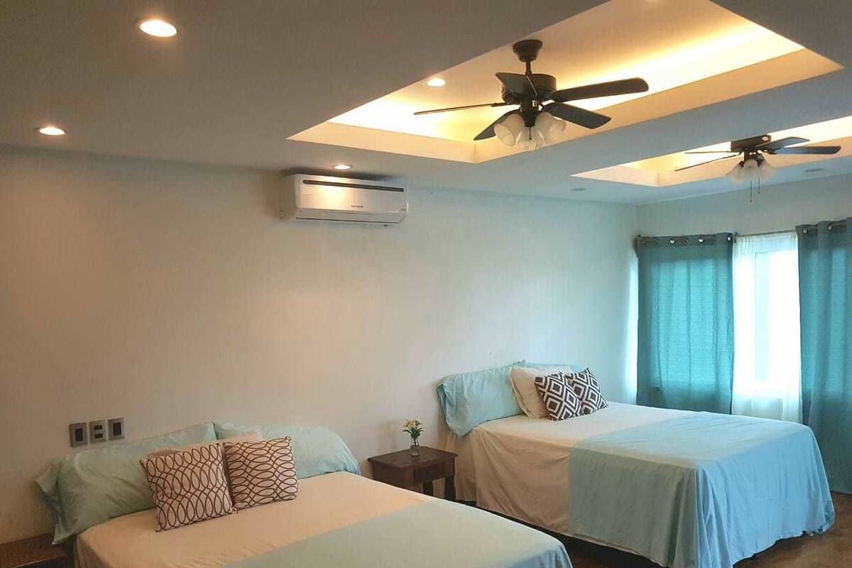 A hotel room with two beds, ceiling fans, an air conditioning unit, and a window with curtains.