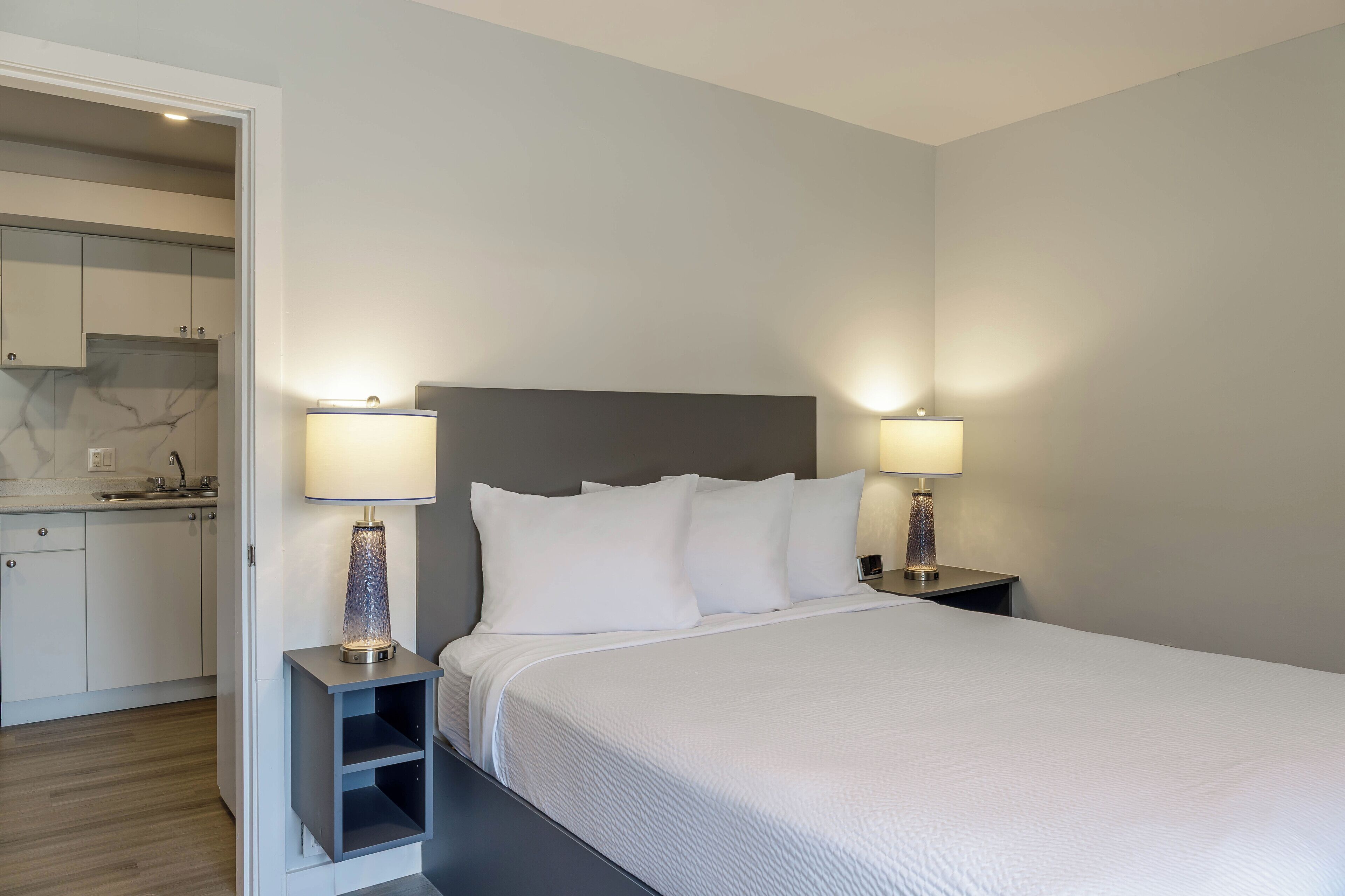 A neatly arranged bedroom with a bed, two bedside tables with lamps, and a view of a kitchen area.