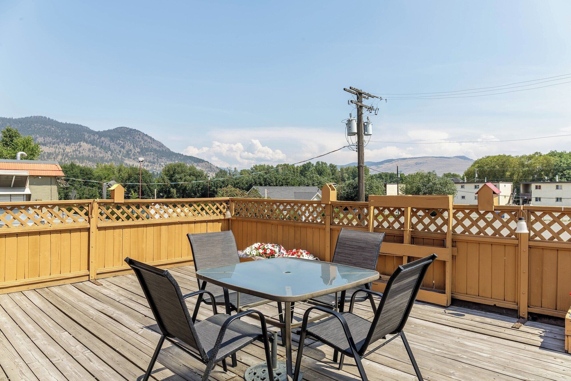 A wooden deck with a glass table and four chairs, surrounded by a wooden fence, overlooking a mountainous landscape.