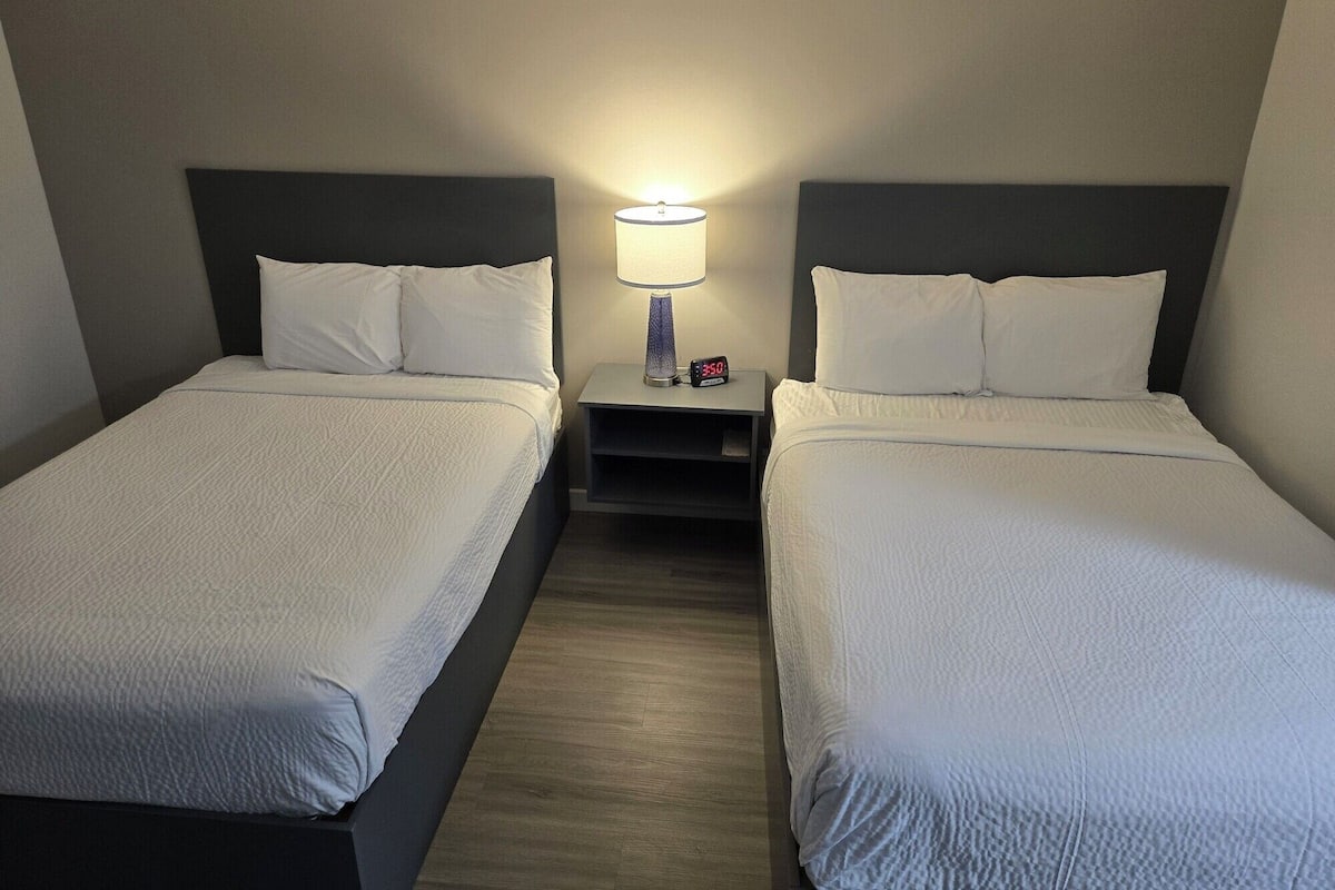 Ein Hotelzimmer mit zwei Einzelbetten, jeweils mit Kopfstütze, Nachttisch und Lampe sowie digitaler Uhr.