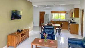 A modern living room with a flat-screen TV, a wooden coffee table with flowers, a blue leather sofa, and a dining area with a table and chairs. - Anse La Mouche Holiday Apartments (Victoria)