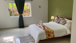 A neatly made bed with a floral bedspread, a bedside lamp, and a window with curtains. - Anse La Mouche Holiday Apartments (Victoria)