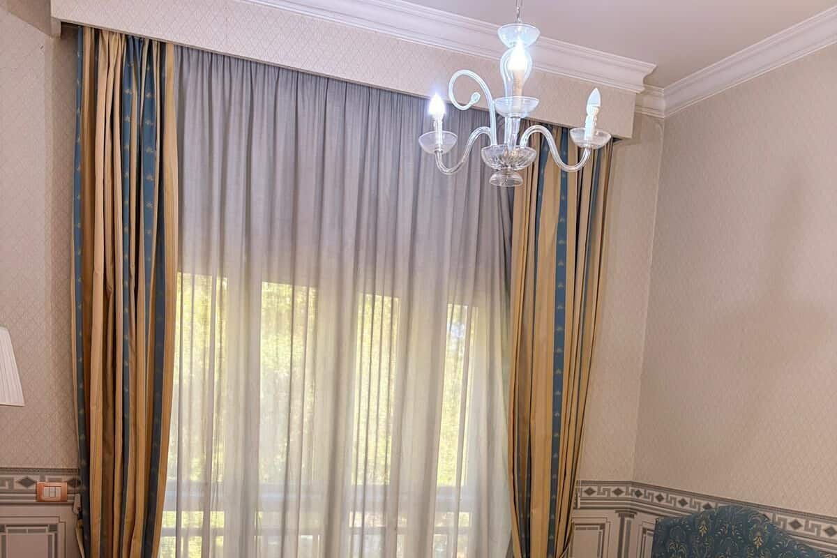 A hotel room with two beds, a wooden desk, a chandelier, and sheer curtains.