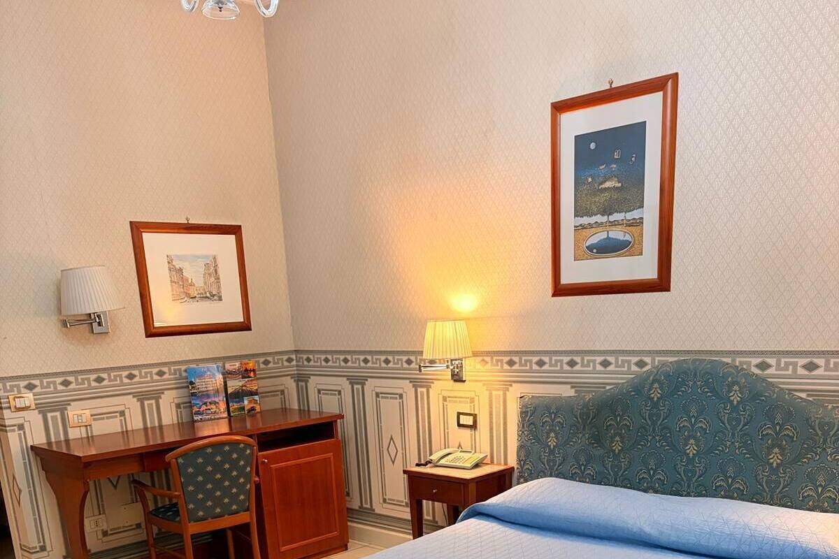 A hotel room with a bed, desk, chair, lamp, and a framed picture on the wall.