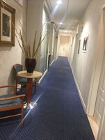 Un pasillo de hotel con alfombra azul, una silla de madera, una mesita pequeña y una planta en maceta.