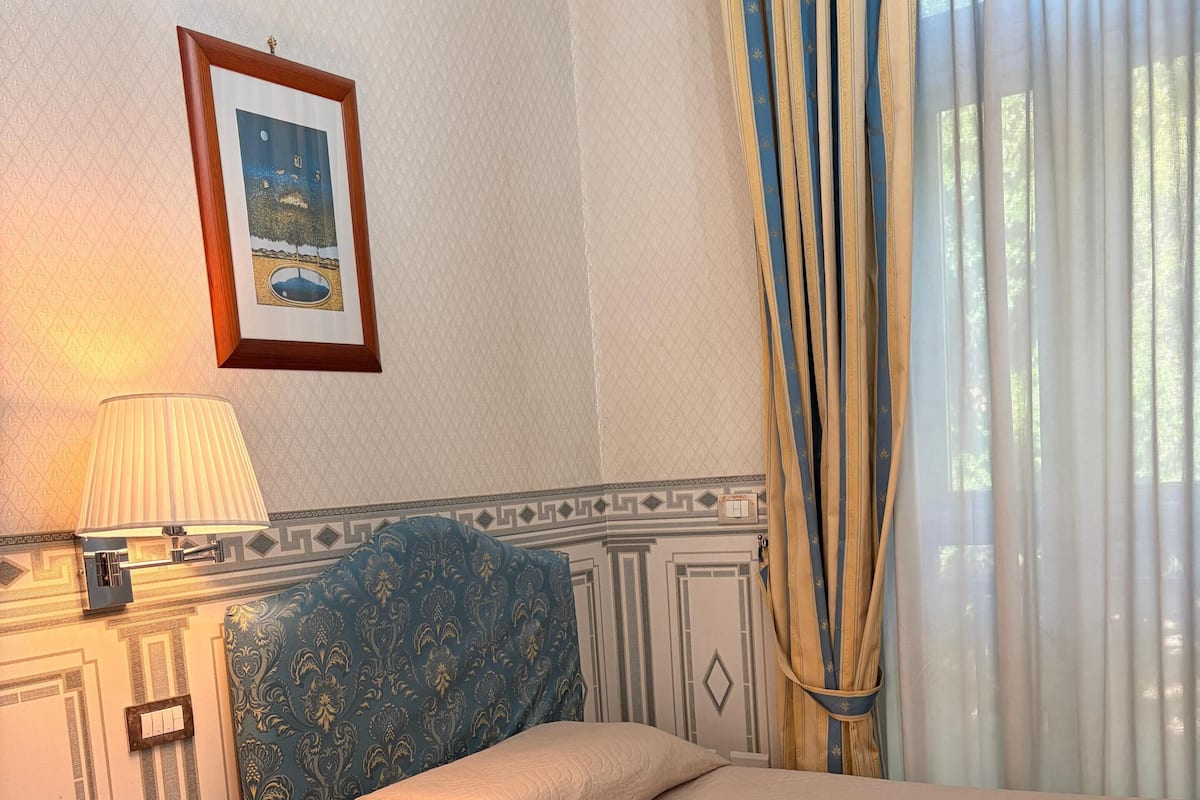 A bedroom with a bed, a bedside table, a lamp, a framed picture on the wall, and a window with curtains.