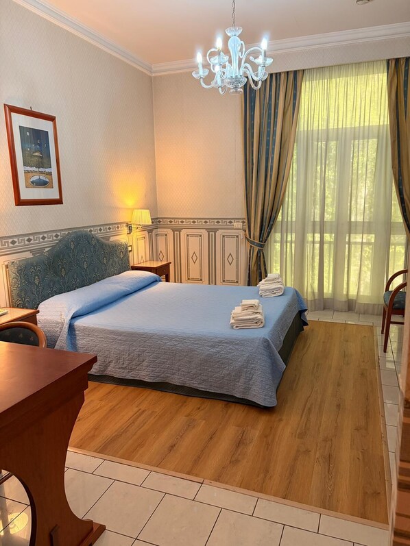 Une chambre d’hôtel avec un lit, un bureau, une chaise, un lustre, une lampe, des rideaux et un tableau encadré au mur.