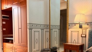 Habitación de hotel con armario de madera, un espejo grande, una mesita de noche, una lámpara, un cesto de basura y toallas ordenadamente dobladas.