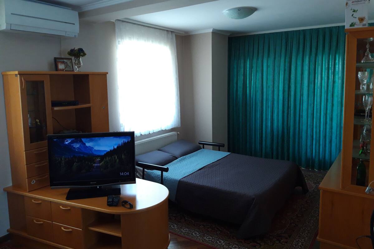 Et hotelværelse med en seng, et fjernsyn på et træstel, et lille bord, et skab og et vindue med gardiner.