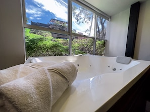 Une salle de bain moderne avec une grande baignoire, une fenêtre donnant sur un paysage verdoyant et des bâtiments, et une serviette posée sur le bord de la baignoire.