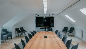 Ein moderner Konferenzraum mit einem großen Holztisch, grauen Stühlen und einem an der Wand befestigten Flachbildfernseher.
