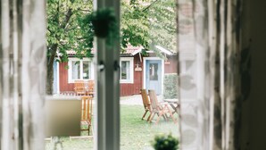 Blick durch ein Fenster. Im Vordergrund ein Holzstuhl und ein kleiner Tisch, im Hintergrund ein Holzzaun und ein Gebäude mit einer blauen Tür.
