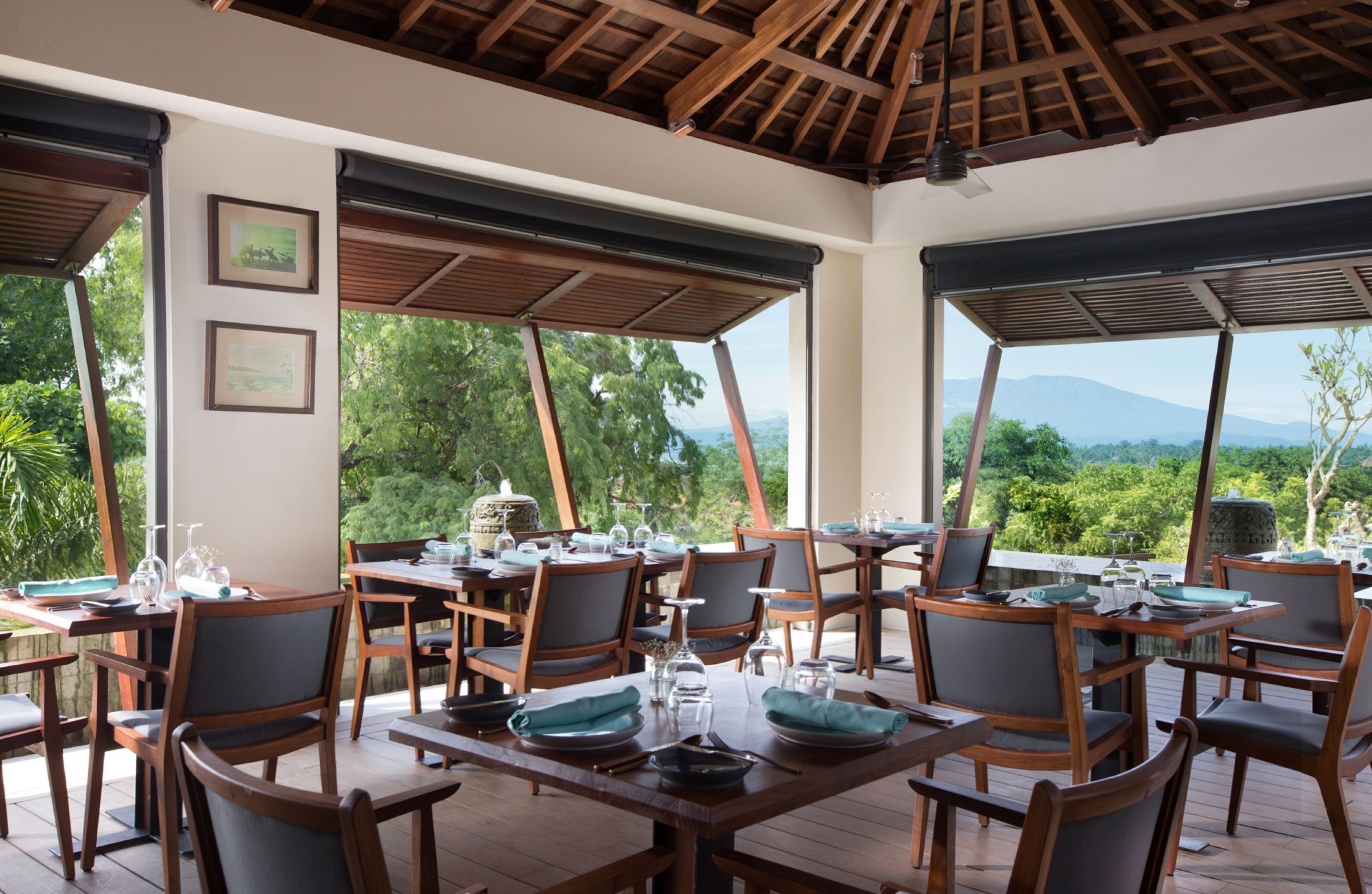 Um restaurante com mesas e cadeiras de madeira, com um cenário de vegetação exuberante e montanhas.