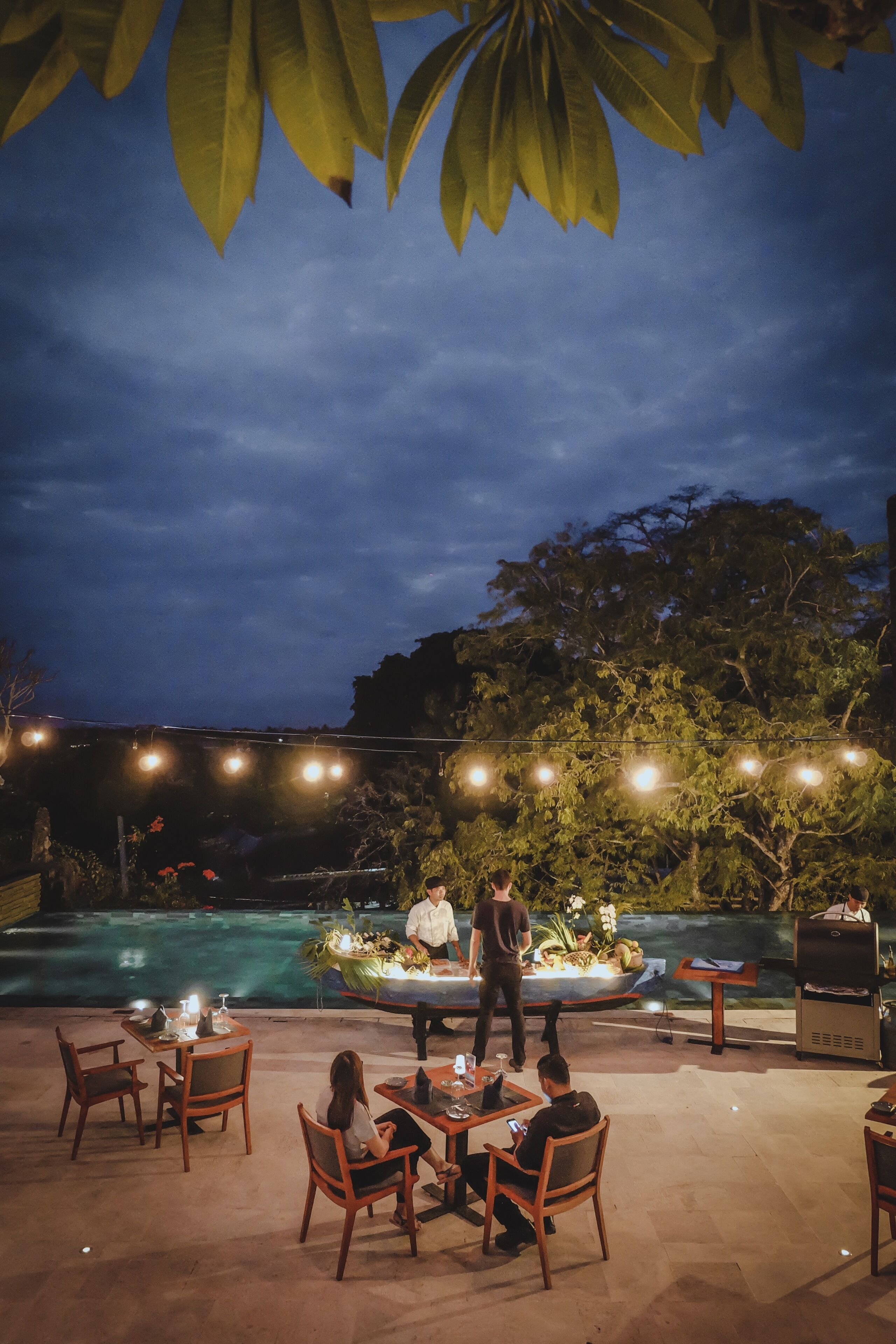 An outdoor dining area with tables and chairs, a pool, and string lights in the background.