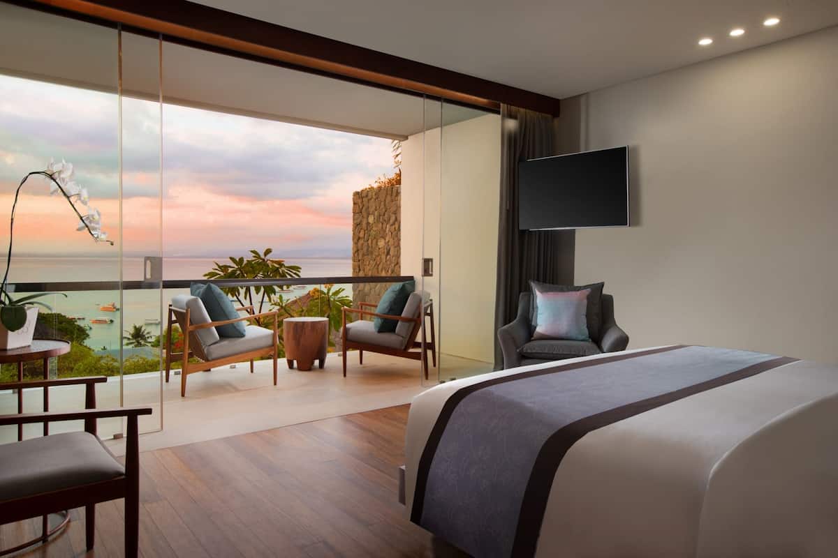Une chambre d’hôtel moderne dotée d’un grand lit, d’un téléviseur fixé au mur et d’un balcon avec coin salon et vue sur l’océan.