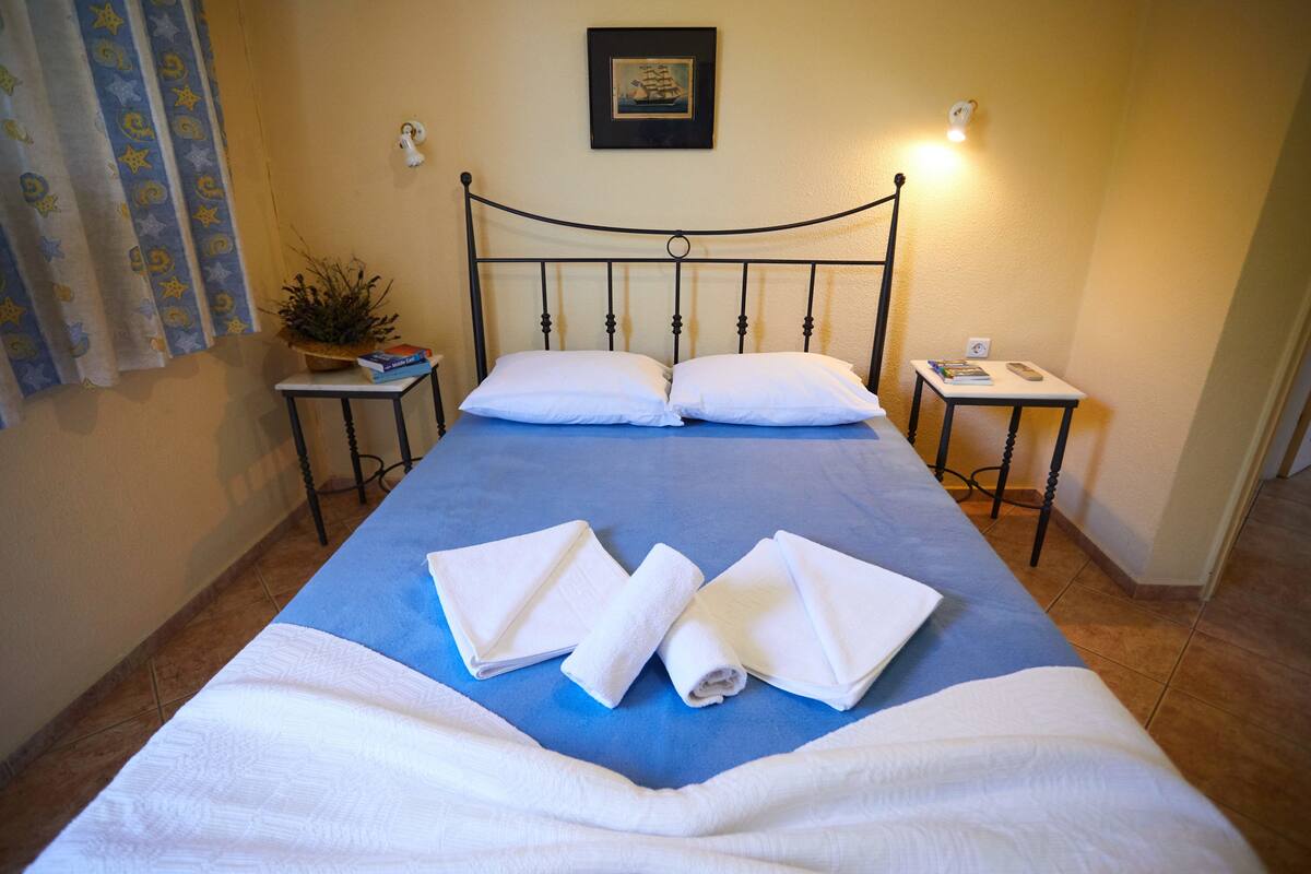 A neatly made bed with white linens and two pillows, a small bedside table with a plant and books, and a wall-mounted picture.