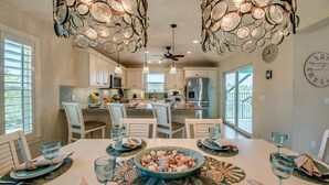 A dining area with a large table set for six, surrounded by white chairs. There is a ceiling fan above and a decorative light fixture with circular elements.