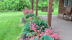Un pabellón de madera con un banco, rodeado de un jardín con plantas verdes grandes y flores rosas.