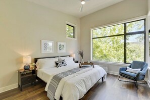 A bedroom with a large window, a bed with a grey and white striped bedspread, a bedside table with a lamp, and a chair.