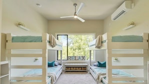 Una habitación con literas, ventilador de techo, equipo de aire acondicionado y ventana con vista a árboles.