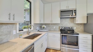 Una cocina moderna con armarios blancos, electrodomésticos de acero inoxidable, un lavamanos doble y una ventana con vistas a zonas verdes.