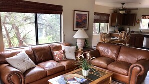 A living room with a brown leather sofa, a wooden coffee table, and a large window with a view of trees.