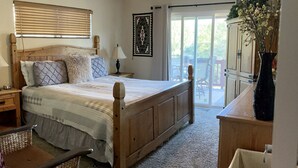 A bedroom with a wooden bed, a ceiling fan, a nightstand, a lamp, a window with blinds, a sliding glass door to a balcony, and a vase with flowers.