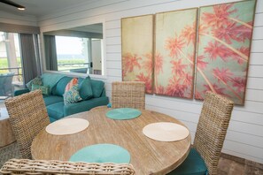 Une pièce avec une table ronde en bois, des chaises en osier, un canapé de couleur turquoise et une grande fenêtre donnant sur un beau paysage.