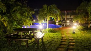 Un espacio al aire libre nocturno con piscina, palmeras y una mesa de picnic.