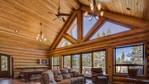 A spacious log cabin interior with a large wooden ceiling, a cozy seating area with a sofa and chairs, a dining table, and large windows offering views of the surrounding forest.
