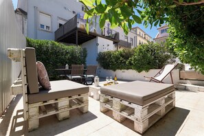 Un área de estar al aire libre con mesas de madera y sillones, rodeada por un muro blanco y vegetación.