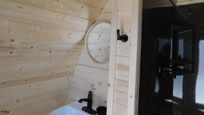Un baño con paneles de madera, un lavamanos blanco, un espejo redondo y un cabezal de ducha negro.