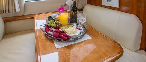 A dining table with a tray of food, including cheese, grapes, and a bottle of wine, in a cabin with leather seating and a window view.