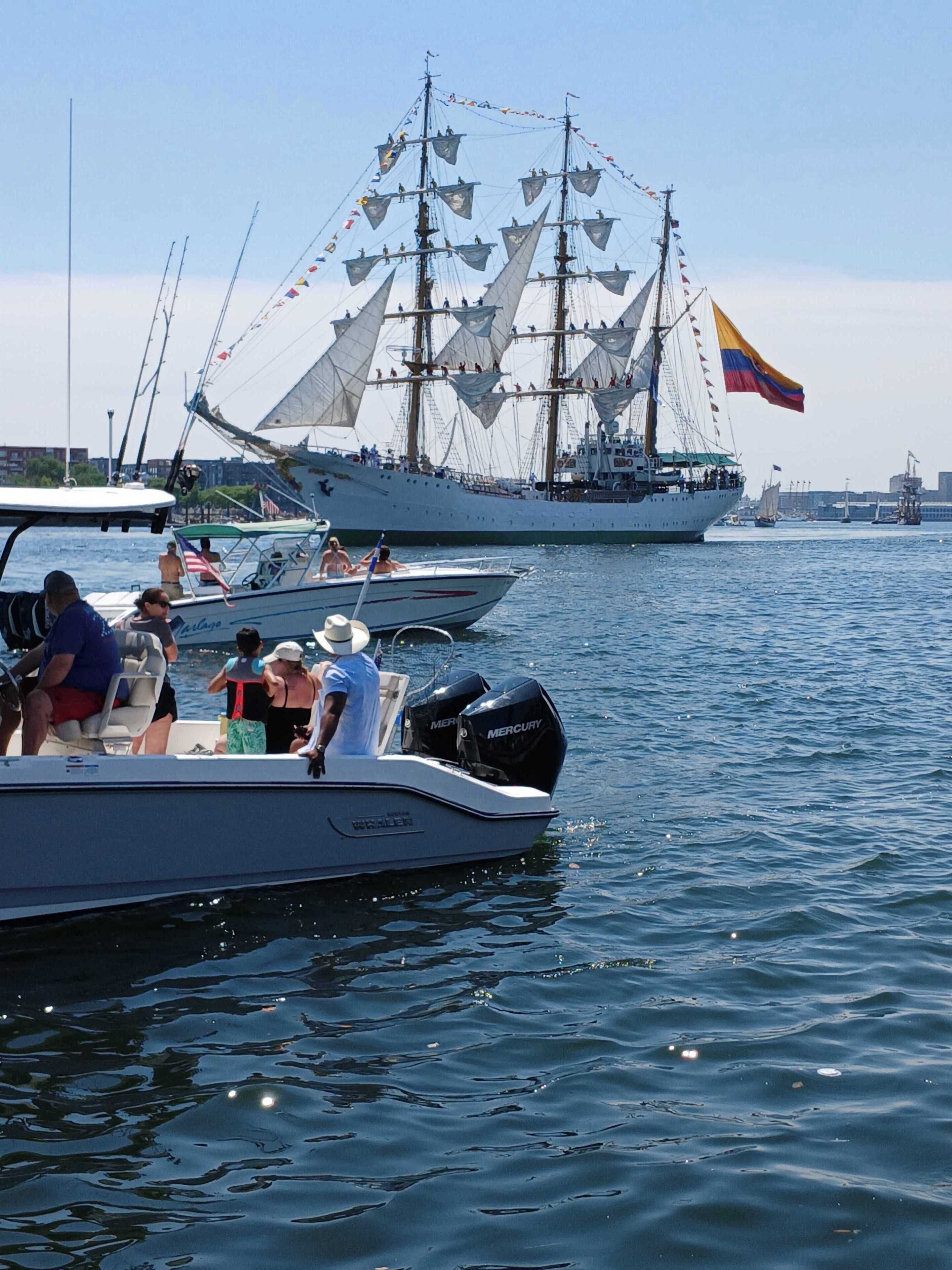 En småbåtshamn med flera båtar, inklusive ett stort segelfartyg med flera master och flaggor, och mindre motorbåtar med folk i.